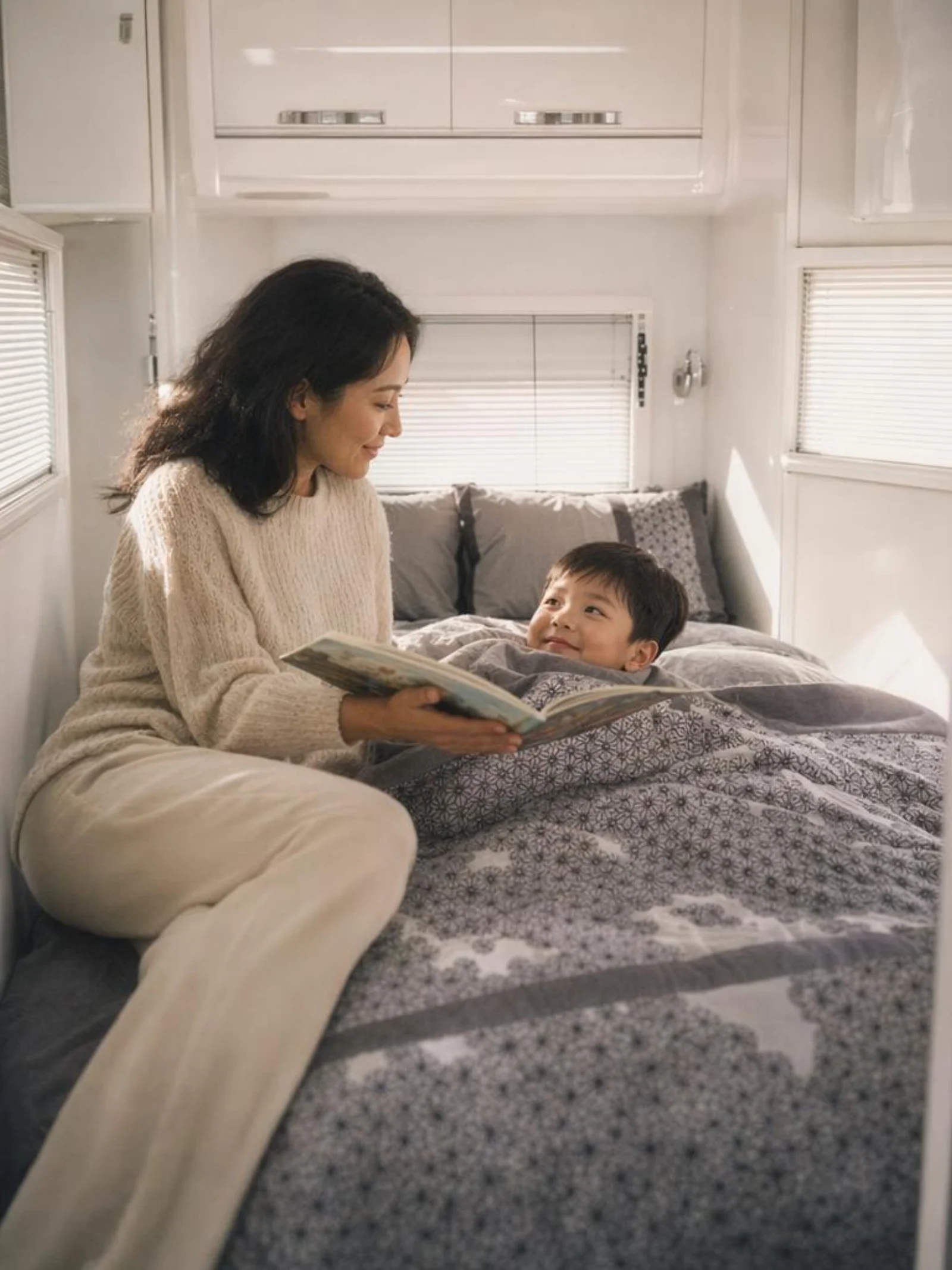 Woman reading a book cross-legged on a queen bed inside a caravan bedroom, soft morning light
